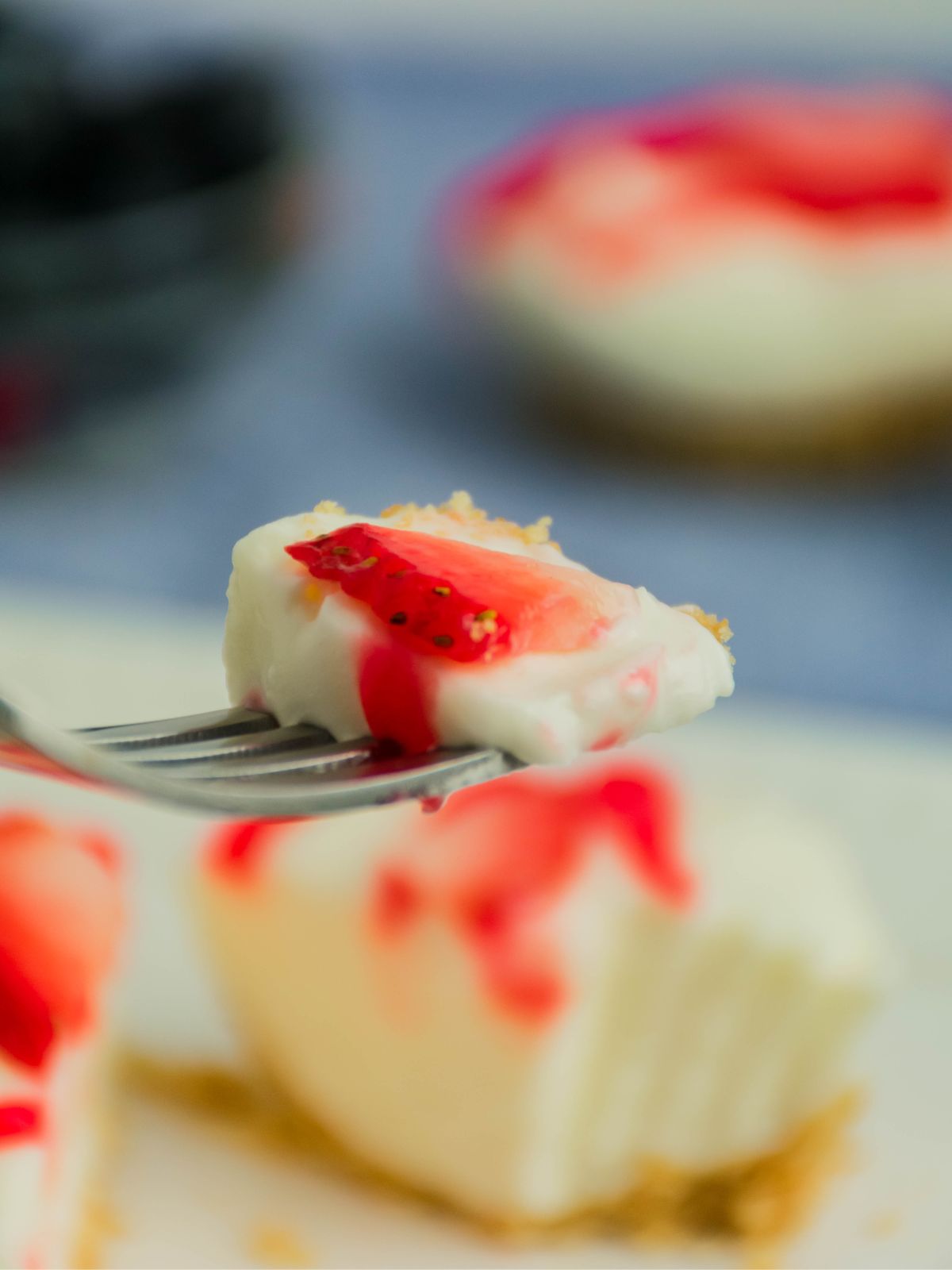 Fork holding a bite of mini cream cheese vanilla pudding dessert topped with strawberry sauce and graham cracker crust in the background.
