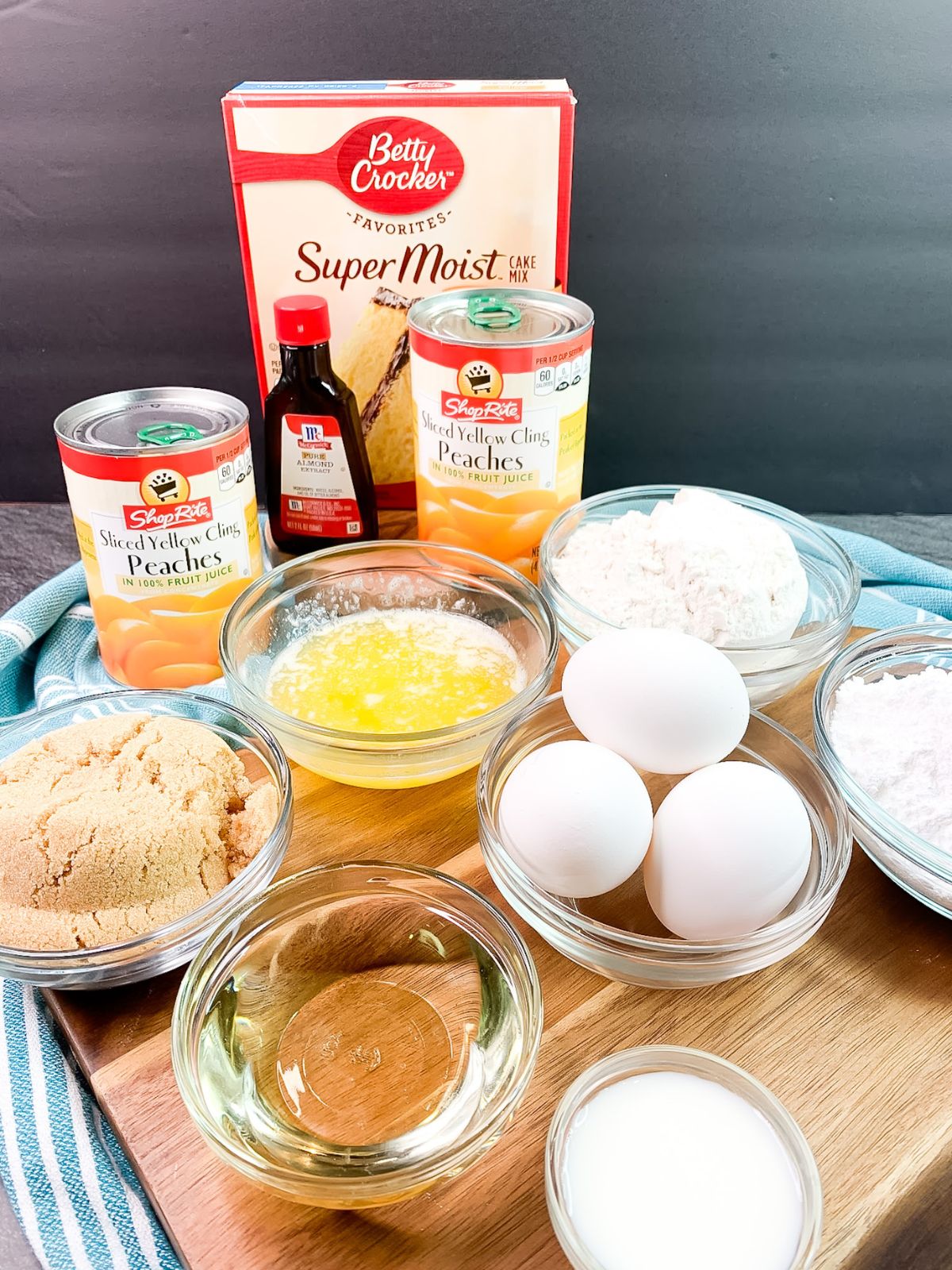 Ingredients for peach crumb cake with yellow cake mix arranged on a wooden board, including canned peaches, yellow cake mix, eggs, almond extract, brown sugar, flour, butter, powdered sugar, milk, and vegetable oil.