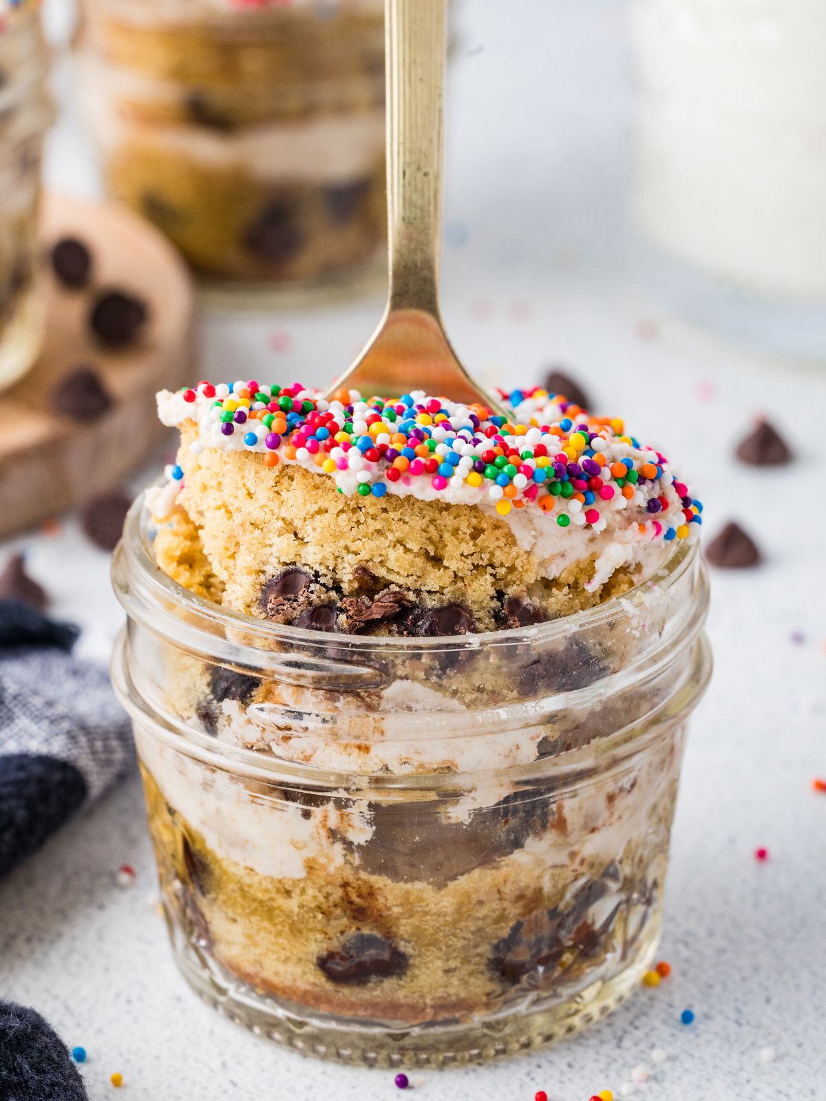Layered chocolate chip cake and frosting in a mason jar topped with rainbow sprinkles and served with a spoon.