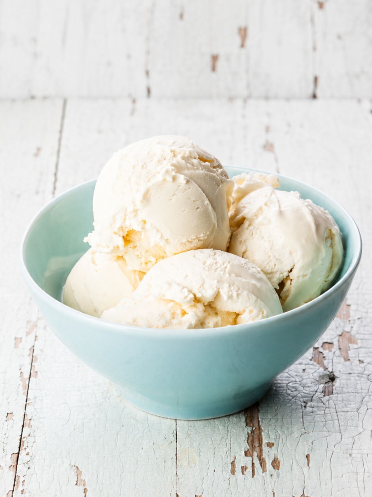 Homemade vanilla ice cream scoops in a light blue bowl on a rustic white wooden table.