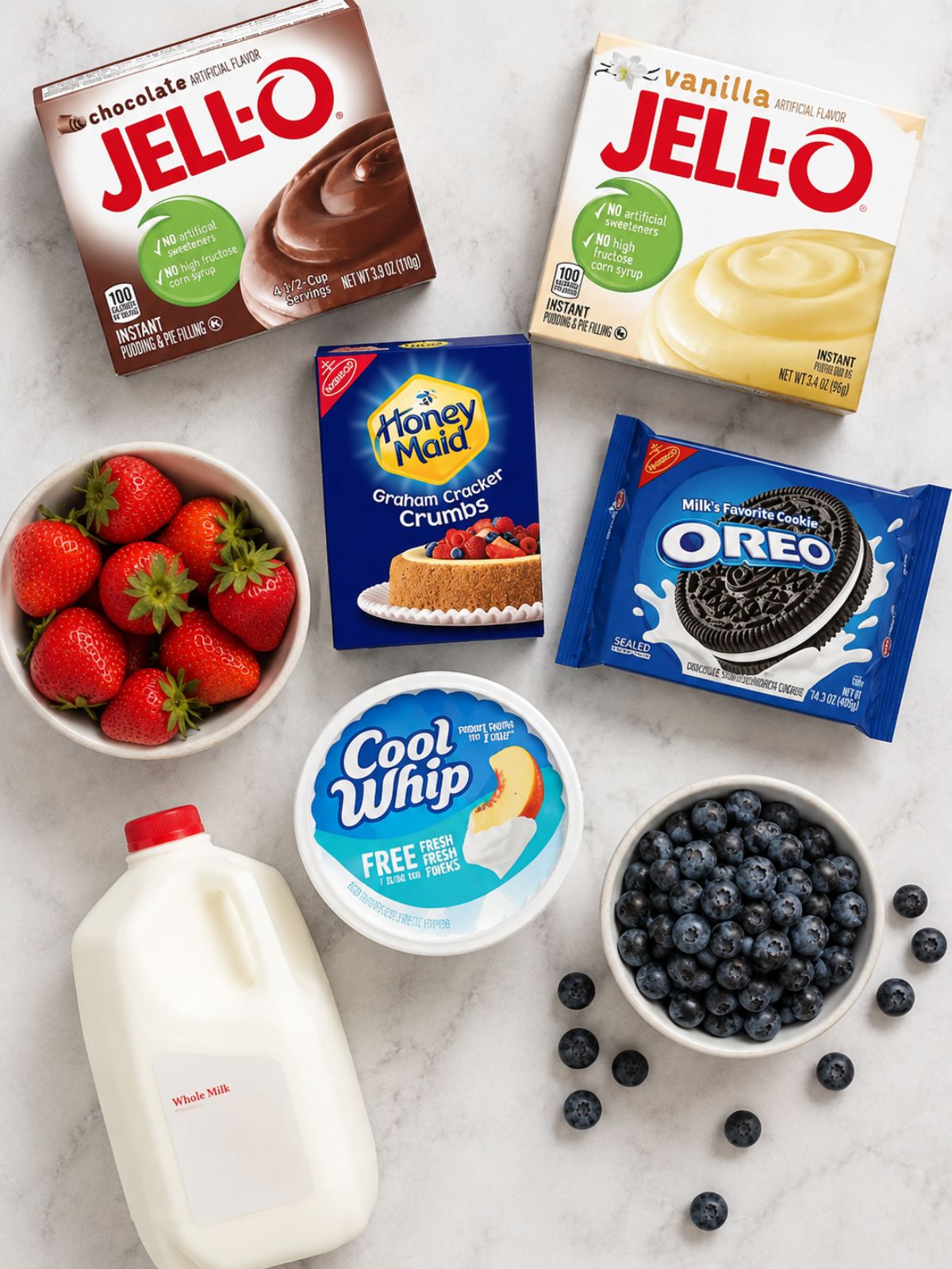 Overhead flat lay of ingredients for mason jar pudding desserts including chocolate and vanilla pudding mix, graham cracker crumbs, Oreo cookies, Cool Whip, milk, fresh strawberries, and blueberries arranged on a marble countertop.