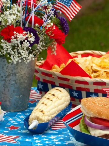 Traditional 4th of July foods featuring grilled corn, a cheeseburger, patriotic table decorations, and festive red, white, and blue picnic accents.