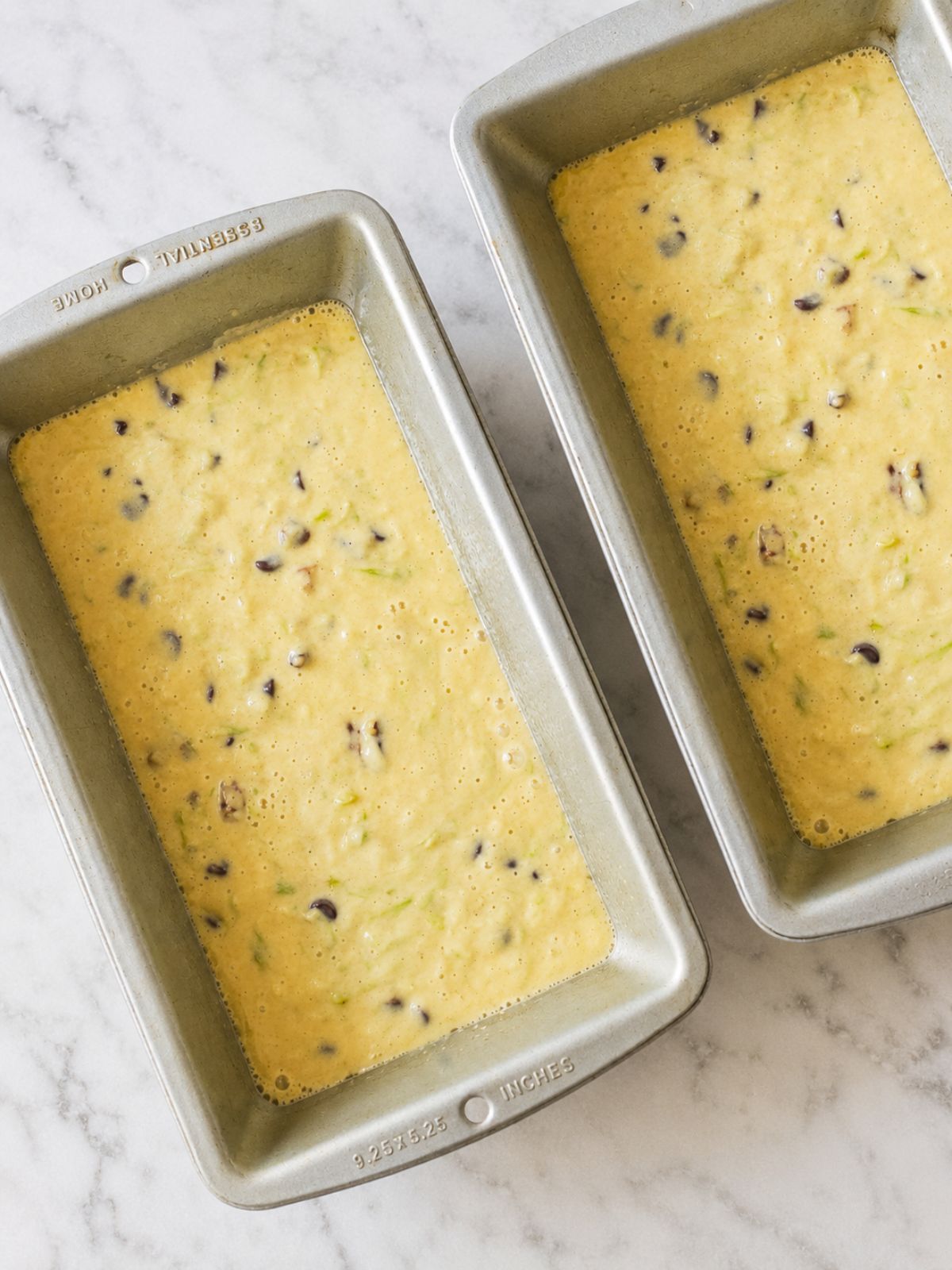 Two loaf pans filled with Bisquick zucchini bread batter mixed with chocolate chips and walnuts before baking on a marble countertop.