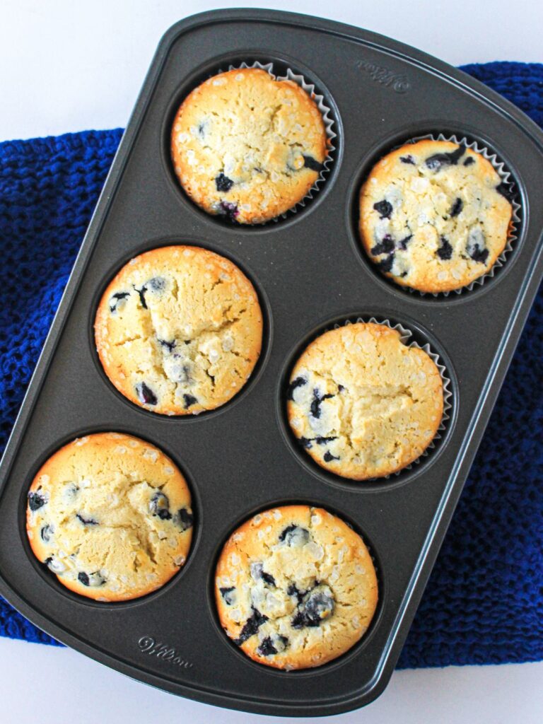 6 baked classic blueberry muffins in baking pan on top of blue napkin.