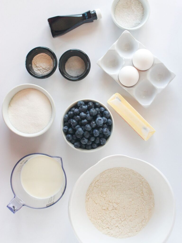 ingredients for classic homemade blueberry muffins in bowls, measuring cups and spoons on white background.