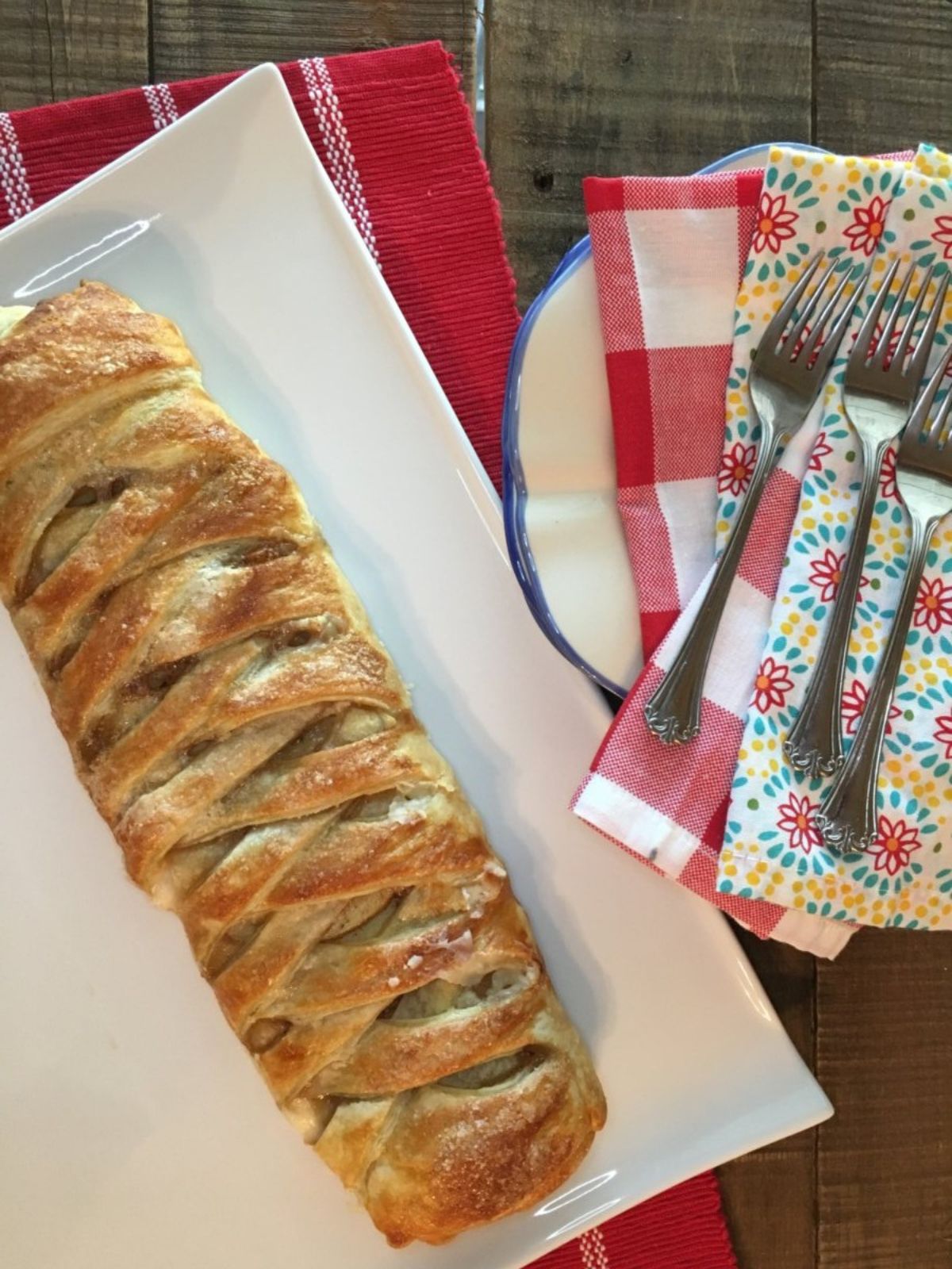 apple braid made with puff pastry on white tray with plate, forks and napkins to the side.