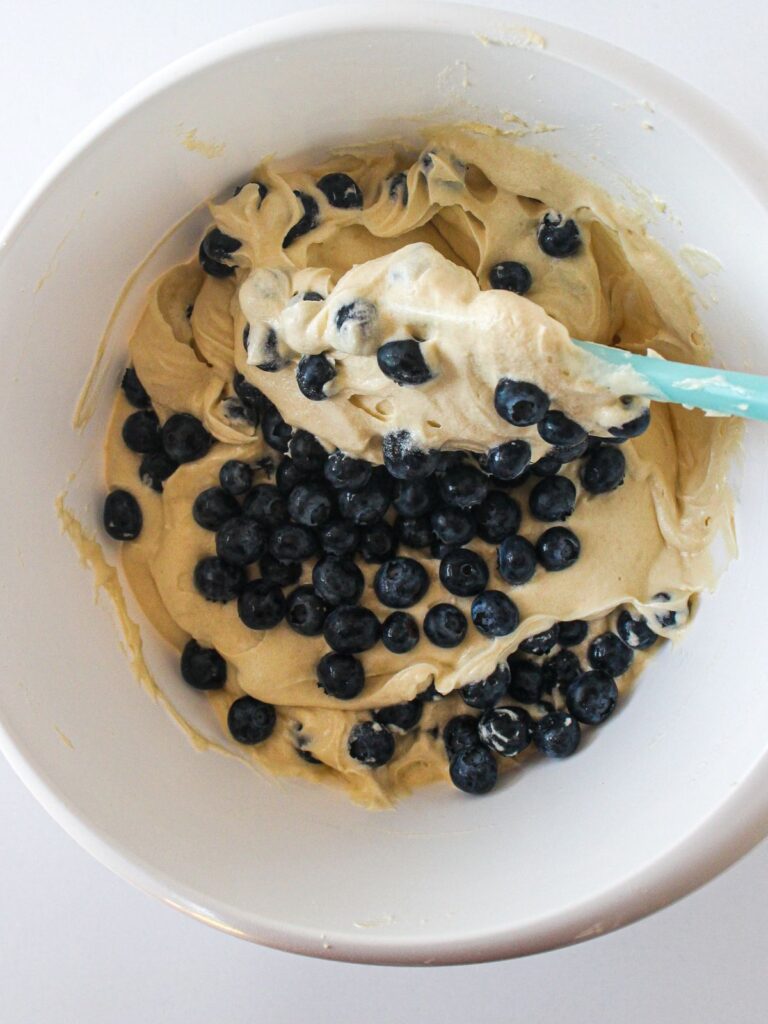 muffin batter in white bowl with blueberries being folded in.